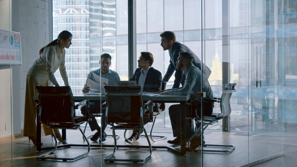 A team of five business professionals meets around a conference table in a modern office setting.